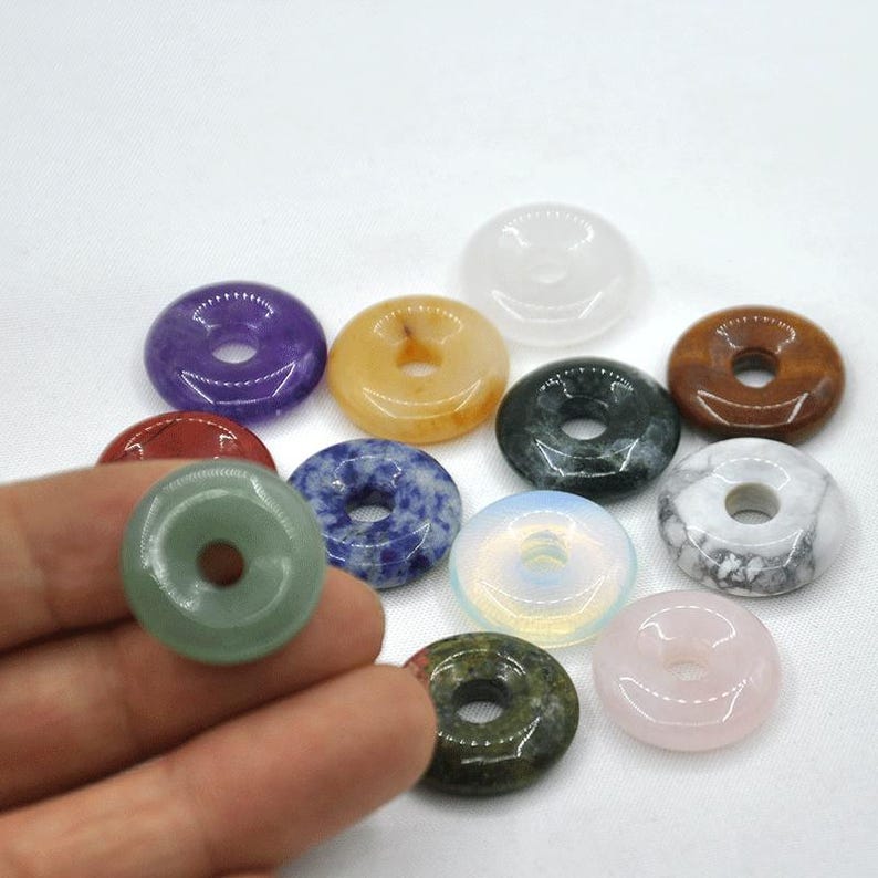 Pode incluir: Conjunto de 10 contas de pedras preciosas em forma de rosquinha em v&aacute;rias cores, incluindo verde, roxo, branco, marrom, azul, laranja e rosa. As contas est&atilde;o dispostas sobre uma superf&iacute;cie branca.