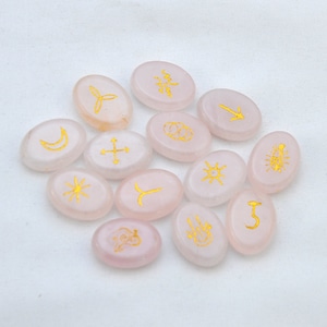 May include: A set of 12 oval-shaped pink rose quartz stones with gold symbols. The symbols include a crescent moon, a sun, a cross, a star, a fish, a wave, a circle, a crescent moon, a trident, a hook, a bow and arrow, and a tree.