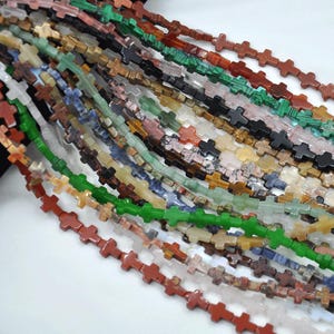 May include: A strand of colorful gemstone cross beads. The beads are made of various stones, including green, red, brown, and white. The cross shape is a popular design for jewelry making.