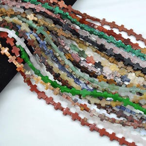 May include: A strand of gemstone beads in various colors, each bead is shaped like a cross. The beads are strung together on a clear thread. The beads are made of natural stone and have a variety of patterns and textures.