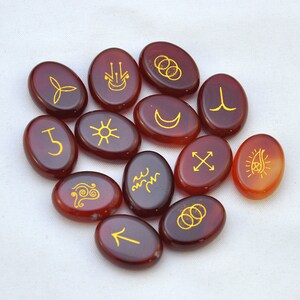 May include: Thirteen smooth, oval-shaped carnelian stones with gold-colored symbols etched on them. The symbols include a crescent moon, a sun, a cross, a wave, a spiral, a trident, a circle with three dots, a circle with a dot in the center, a circle with a dot in the center and a line through it, a circle with a dot in the center and a line through it, a circle with a dot in the center and a line through it, a circle with a dot in the center and a line through it, and a circle with a dot in the center and a line through it.