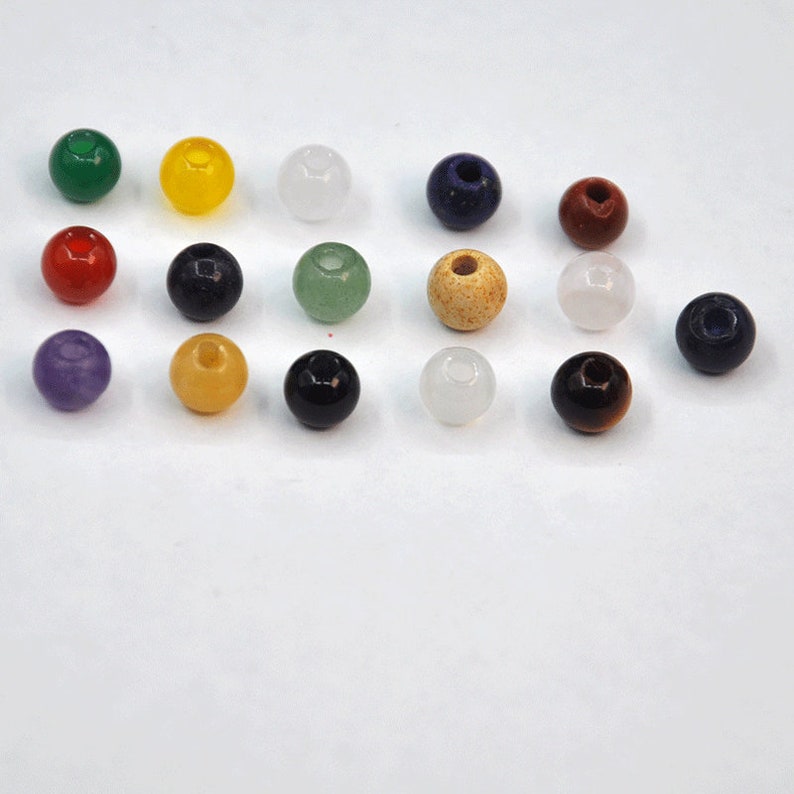 May include: Assortment of 15 round gemstone beads in various colors, including green, yellow, red, purple, blue, brown, black, and white. The beads are arranged in rows on a white surface.