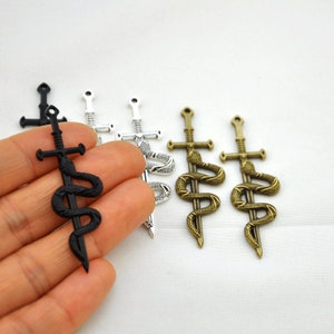 May include: Three sword and snake charms in black, silver, and bronze. The charms are detailed with a snake wrapped around a sword.