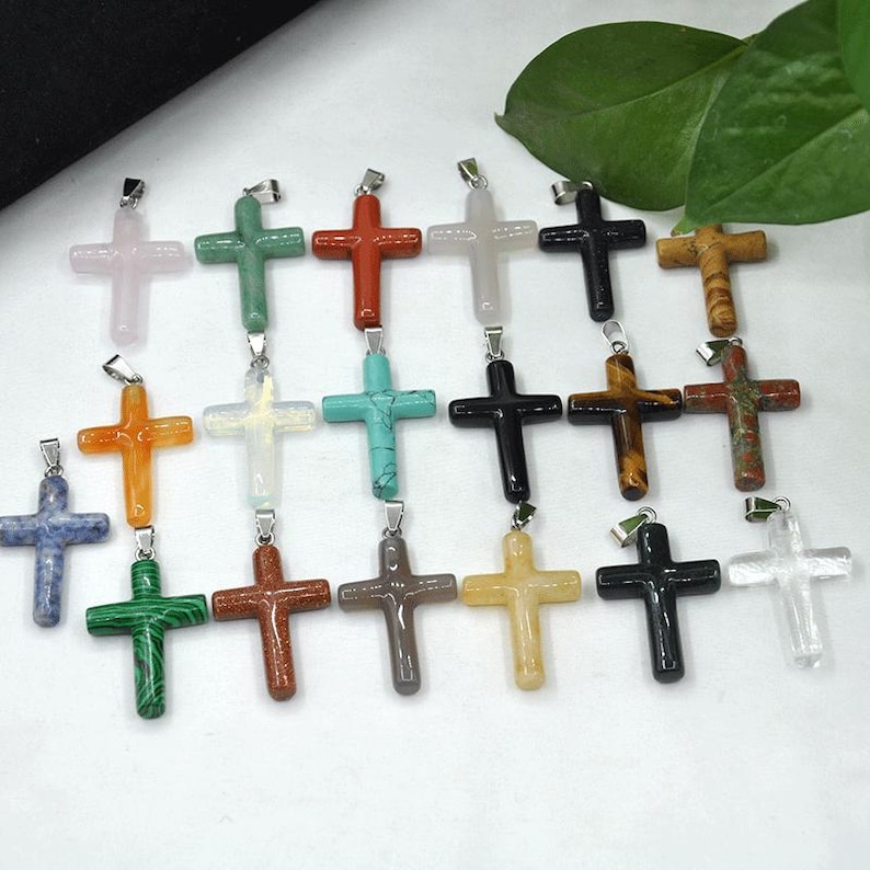 May include: A selection of gemstone cross pendants in a variety of colours, such as pink, green, red, white, black, and brown. Each cross features a silver-coloured bail for attaching to a chain. The crosses are made from different types of stone.