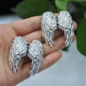 May include: Three silver-colored wing charms with detailed feather textures. Each wing has a small loop at the top for attaching to jewelry or other crafts. The wings are displayed against a blurred background.