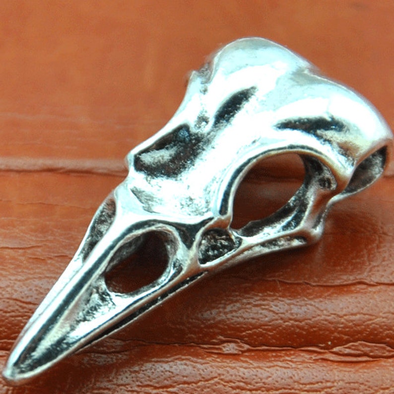 May include: Silver-toned bird skull pendant with a detailed, intricate design. The pendant has a hollowed-out eye socket and beak.