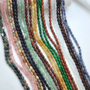 Pu&ograve; includere: Assortimento di perline di pietre preziose di forma ovale in vari colori, tra cui verde, blu, rosa, marrone e bianco. Le perline sono infilate su uno sfondo bianco.