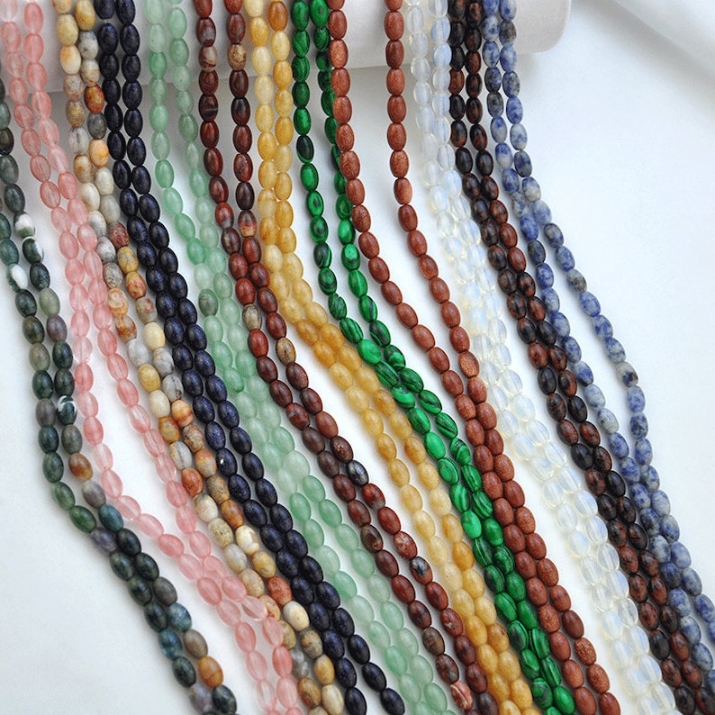 Pu&ograve; includere: Assortimento di perline di pietre preziose di forma ovale in vari colori, tra cui verde, blu, rosa, marrone e bianco. Le perline sono infilate su uno sfondo bianco.