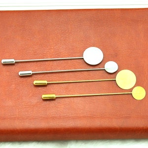May include: Four different sized, round, blank metal pins with a silver and gold finish. The pins are on a brown leather surface.