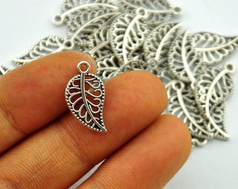 Op de afbeelding: Zilverkleurige blad-vormige bedels met ingewikkelde openwerk ontwerpen. De bedels hebben een kleine lus aan de bovenkant om aan sieraden of andere knutselwerken te bevestigen. Een hand houdt een bedel vast, met nog veel meer op de achtergrond.