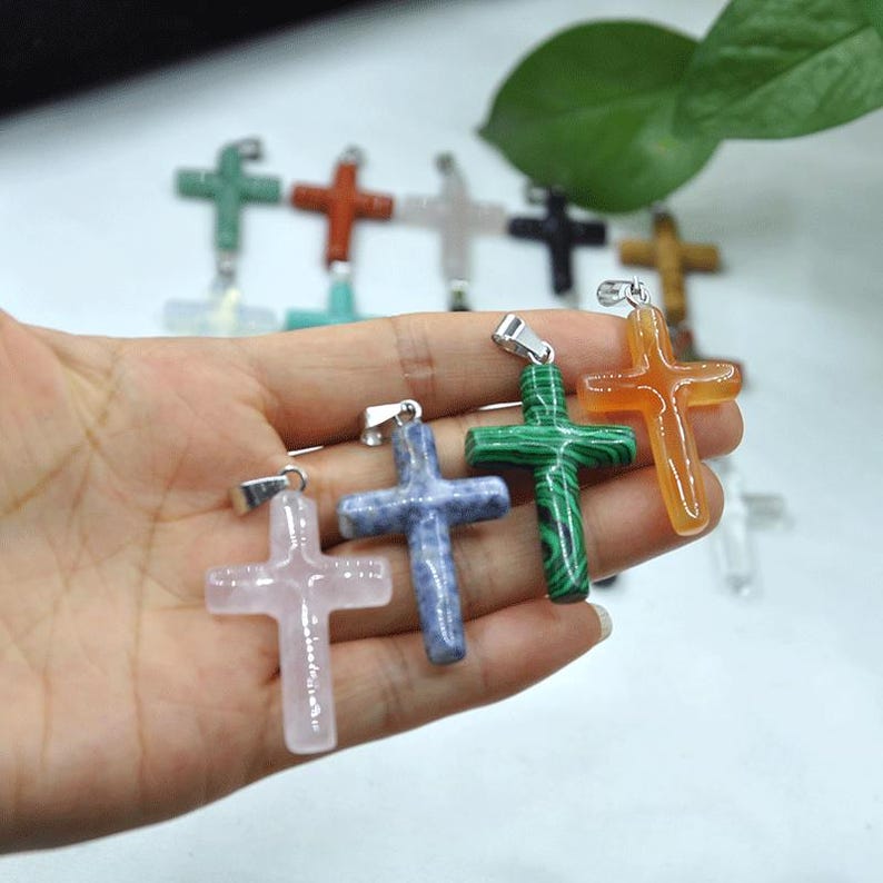 May include: A selection of gemstone cross pendants in shades of pink, blue, green, and orange. Each cross features a silver-coloured bail for attaching to a chain. The crosses are crafted from polished stone.