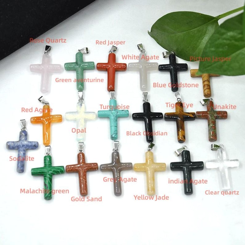 May include: A selection of gemstone cross pendants in various colours and materials, including rose quartz, red jasper, and turquoise. Each cross has a silver-tone bail for hanging. The image includes labels identifying each stone type.