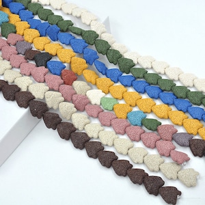 Pode incluir: Um fio de contas de rocha vulcânica em várias cores, incluindo marrom, branco, verde, azul, amarelo e rosa. As contas têm o formato de coração e são enfiadas em um fio transparente.