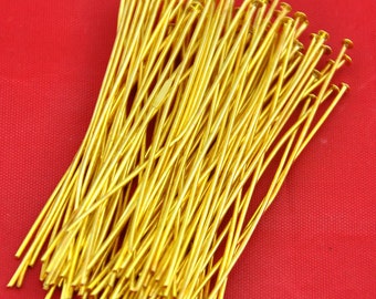 May include: A large collection of long, thin, gold-colored metal headpins, each featuring a small, flat, circular head at one end and a straight, slender shaft, arranged in a dense, overlapping cluster against a solid red background.