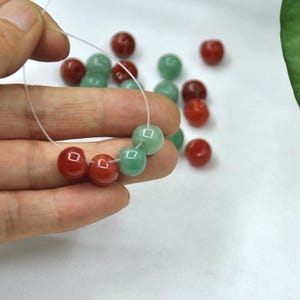 Puede incluir: Un hilo de cuentas redondas rojas y verdes. Las cuentas están ensartadas en un cordón fino y transparente. Cuentas sueltas adicionales de los mismos colores están dispersas en el fondo. Las cuentas tienen un acabado brillante.