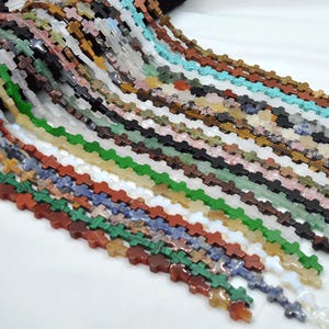 May include: A variety of cross-shaped gemstone beads in different colors and patterns. The beads are strung together in long strands, creating a colorful and textured display.