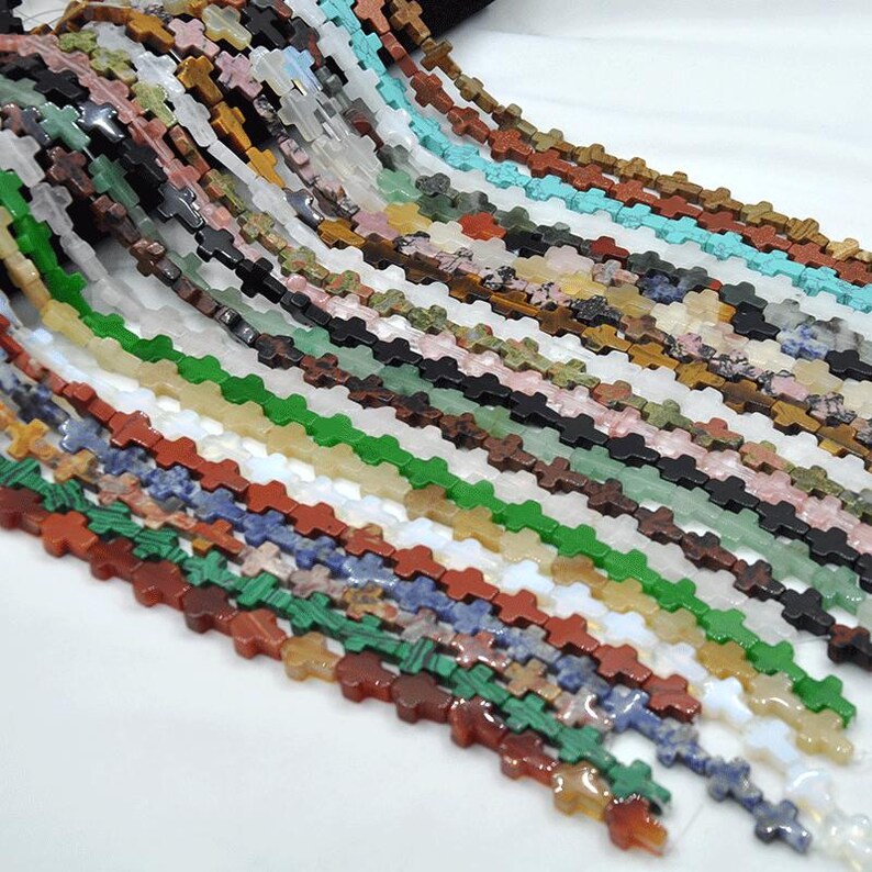 May include: A variety of cross-shaped gemstone beads in different colors and patterns. The beads are strung together in long strands, creating a colorful and textured display.