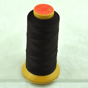 May include: A spool of black thread, tightly wound around a yellow core. The top of the spool is orange with gold text and a small illustration. The thread is a solid, dark color, suitable for various sewing projects.