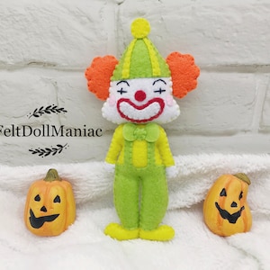 Può includere: Un pupazzo di clown di feltro con un vestito verde e giallo, capelli arancioni e un naso rosso. Il clown ha un sorriso dipinto e occhi a forma di croce. Il pupazzo è in piedi davanti a due zucche arancioni a forma di jack-o'-lantern.
