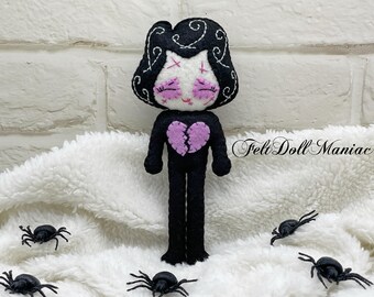 May include: A handmade felt doll with black hair and a black outfit, featuring a purple broken heart design. The doll has closed eyes and a white face with pink accents. Small black spiders are scattered around the doll on a white, fluffy surface. The text "Felt Doll Maniac" is visible.