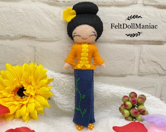 May include: A felt doll with black hair in a bun, wearing a yellow top with a blue skirt and a yellow flower on her head. The doll is standing on a white surface with a brick wall in the background. The text "FeltDollManiac" is visible in the top right corner of the image.