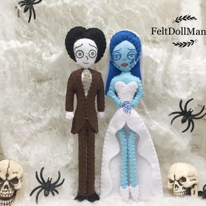 May include: Felt doll couple dressed as a bride and groom, inspired by the characters from the movie Corpse Bride. The bride is wearing a white dress with a blue bodice and a blue veil. The groom is wearing a brown suit with a white shirt and a black bow tie. The dolls are standing on a white background with a spiderweb pattern.