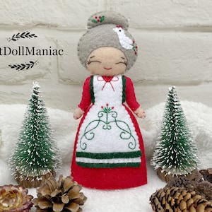 May include: A felt Christmas doll with a red dress, white apron, and gray hair. The doll is holding a small white bird in its hand. The doll is standing on a white surface with two small green trees and pine cones.
