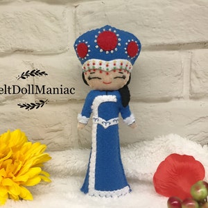 Könnte beinhalten: Filzpuppe in einem blauen Kleid und einer rot-weißen Krone. Die Puppe hat schwarze Haare und ein lächelndes Gesicht. Die Puppe steht auf einer weißen Oberfläche.