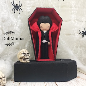 May include: A felt vampire doll lying in a red-lined coffin with a black felt cape. The coffin is on a black felt base. The doll has a black felt suit, white shirt, and black hair. The doll has a small smile and fangs.