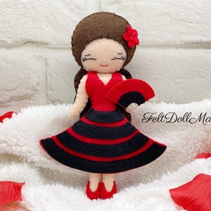 Pode incluir: Uma boneca de feltro vestida com um vestido de flamenco vermelho e preto, segurando um leque preto. A boneca tem cabelo castanho e uma flor vermelha no cabelo. "Felt Doll Maniac" está escrito no fundo branco.