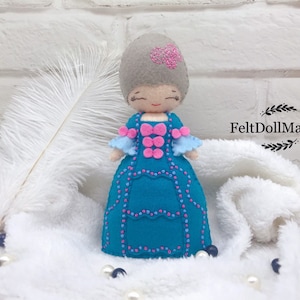 May include: A felt doll wearing a blue dress with pink and white accents. The doll has gray hair and a pink flower on her head. The doll is sitting on a white fluffy surface. FeltDollManiac is written in black text on a white background.