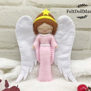 May include: A felt angel ornament with white wings, a pink dress, a yellow star crown, and brown hair. The angel is standing on a white fluffy surface.