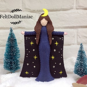May include: Felt doll with a blue dress and a black cape with white stars and a yellow crescent moon on her head. The doll is standing in front of a white brick wall with two small green trees on either side.