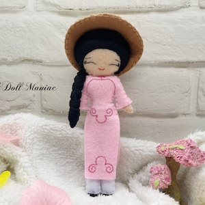 Peut inclure: Une poupée en feutre rose portant une robe rose et un chapeau brun. La poupée a les cheveux noirs en tresse et des détails brodés sur la robe. La poupée est debout sur une surface blanche et moelleuse.