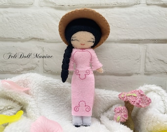 May include: A pink felt doll wearing a pink dress and a brown hat. The doll has black hair in a braid and embroidered details on the dress. The doll is standing on a white fluffy surface.