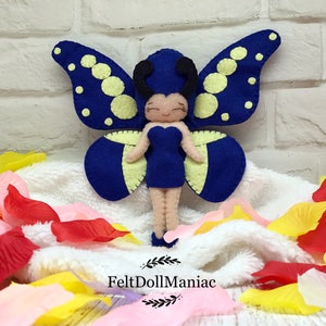 May include: A blue felt butterfly fairy doll with yellow accents and black eyes. The doll is standing on a white fluffy surface with pink and yellow flower petals.
