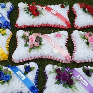 May include: Ten white pillow arrangements with colourful flowers and personalised ribbons. The ribbons have names written on them, such as "Cousin", "Nan", and "Barbara".