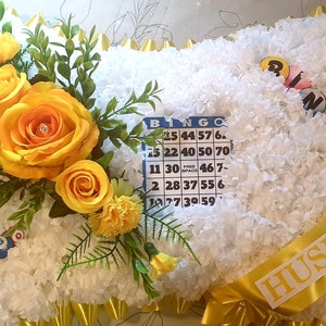 May include: A white pillow shaped floral arrangement with yellow roses and a bingo card. The pillow has a yellow ribbon with the word "HUSBAND" written on it.