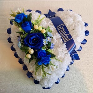 May include: A white heart-shaped funeral wreath with blue carnations and roses. A blue ribbon with the word "Grandad" is tied around the wreath.