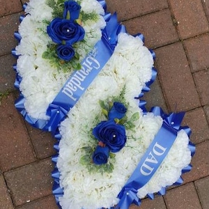 May include: Two white heart-shaped flower arrangements with blue ribbon and blue roses. The ribbons have the words "Grandma" and "Dad" written on them.