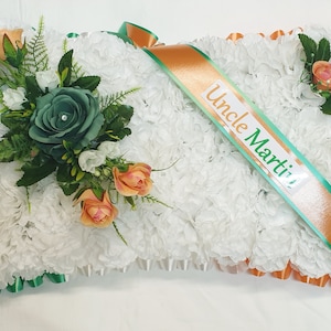 May include: A white pillow with a green and orange ribbon. The ribbon has the text "Uncle Martin!" on it. The pillow is decorated with white carnations, green roses, and orange roses.