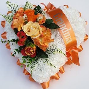 May include: Orange and white heart-shaped funeral arrangement with a satin ribbon that says "Grandma".