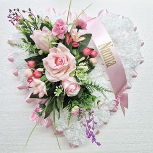 May include: A white heart-shaped funeral wreath with pink roses, pink carnations, and purple flowers. A pink ribbon with the word "Nanna" in gold lettering is tied around the wreath.