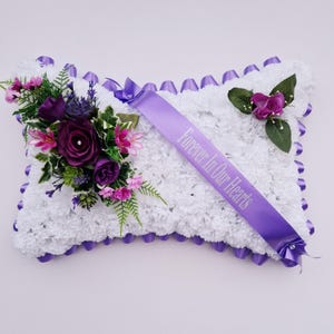 May include: A white pillow with a purple satin trim and a purple ribbon with the text "Forever In Our Hearts" in white lettering. The pillow is decorated with a bouquet of purple and pink flowers.