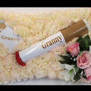 May include: A white pillow with pink flowers and a white cigarette with the word "Granny" on it. The cigarette is decorated with a red and gold band. The pillow is surrounded by pink flowers.