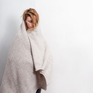 May include: A woman wearing grey jeans is wrapped in a large, light grey blanket. She is looking down and her hair is covering her face.
