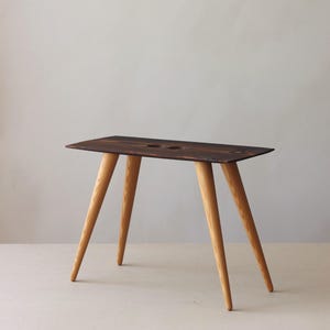 以下が含まれることがあります： 濃い茶色の天板と、明るい色の斜めになった3本の脚を持つ、小さく長方形の木製テーブル。 天板には2つの円形の切り抜きがあります。 テーブルは装飾用または機能的な使用を目的としている可能性があります。