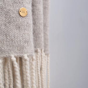 Close-up of a beige knitted wool scarf with off-white fringe and a custom engraved brass monogram pin.