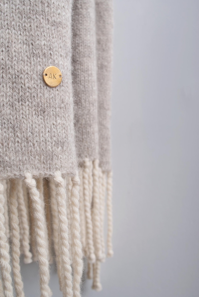 Close-up of a beige knitted wool scarf with off-white fringe and a custom engraved brass monogram pin.