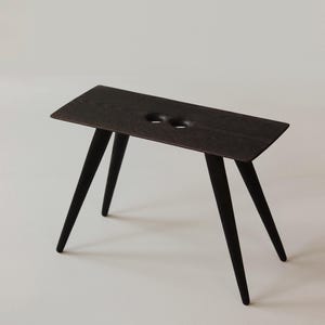 May include: A dark brown wooden stool with a rectangular seat and four angled legs. The seat has two circular cutouts. The stool is made of wood and has a minimalist design.